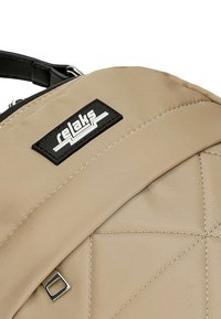 Sac à dos beige avec tissu texturé, présentant des coutures diagonales, un patch logo noir et un détail en métal carré argenté.