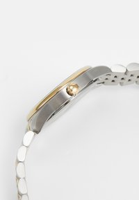 Relógio de metal em tom prateado e dourado com caixa redonda, bezel liso e design de bracelete ligado. Possui uma coroa gravada com o logótipo e acabamento polido.