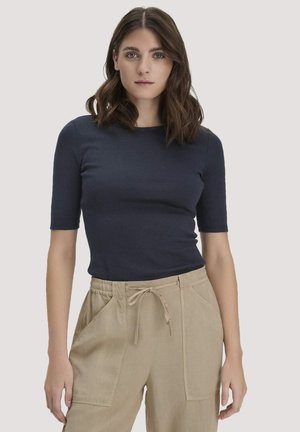 Frau mit schulterlangem braunem Haar, die ein marineblaues halbärmeliges Oberteil und beige Hose mit Kordelzug trägt, vor einem schlichten Hintergrund stehend.