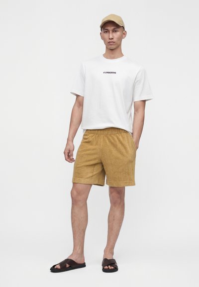 Homme portant une casquette beige, un t-shirt blanc avec logo, un short moutarde texturé et des sandales marron foncé slip-on, debout devant un fond uni.