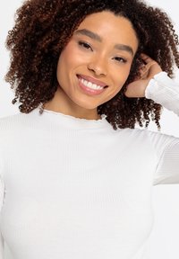 Witte geribbelde langemouwen top met een gekruide halslijn. De stof is zacht, met een aansluitende pasvorm, en het model glimlacht terwijl ze haar haar vasthoudt.