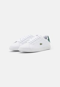 Un paio di sneakers bianche in pelle con lacci, tasselli verdi sul tallone e piccolo logo di coccodrillo verde sui lati, su uno sfondo bianco.