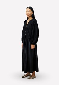 Frau steht, trägt eine schwarze Bluse mit Knöpfen und Puffärmeln, einen langen schwarzen Rock und flache Schuhe aus metallic-gold.