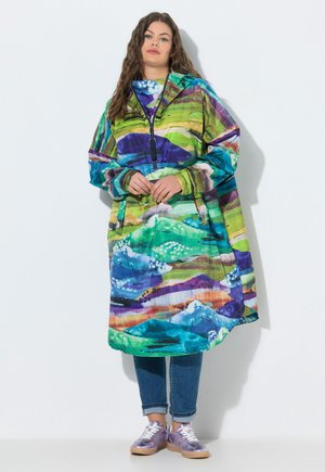 Ung kvinde med langt krøllet hår står iført en flerfarvet oversized hættetrøjekjole over opkørte jeans og lilla sneakers.