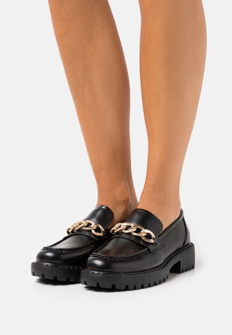 Des mocassins noirs épais avec des semelles épaisses et un détail en chaîne dorée portés pieds nus sur un fond blanc.