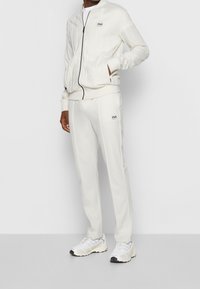 Witte trainingspak met een zip-up jack en bijpassende broek. Kenmerkt zich door ribbelstructuur en logo-accenten. Gepaard met witte sportschoenen.