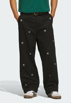 Person wearing black wide-leg pants with small white skull and crossbones patterns, white sneakers with black stripes, and a tucked green shirt.