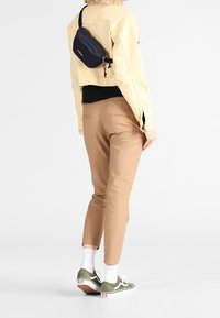 Beige kort jacka, svart topp, tan byxor, gröna och vita sneakers, samt en liten svart midjeväska med axelrem.