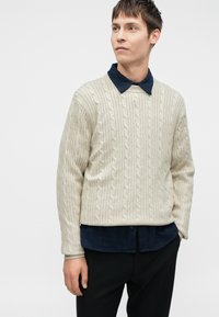 Pull en maille beige fait de tissu texturé, superposé sur une chemise à col bleu foncé. Le pull présente des manches longues et un col rond.