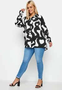 Camicia a righe in bianco e nero con un design astratto e fluido, dalla vestibilità comoda, con chiusura a bottoni e maniche lunghe, abbinata a jeans skinny blu.