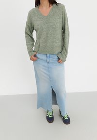 Pull à manches longues vert olive avec un ourlet côtelé, associé à une jupe en denim bleu clair, longueur genou et avec une fente avant, portée avec des baskets bleu marine.