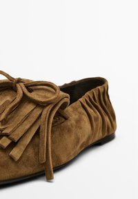 Braune Wildleder-Flatschuhe mit Fransenverzierung und gebundenem Akzent. Die Rückseite weist gerafftes Material für eine weichere Passform auf. Flache Sohlenkonstruktion.