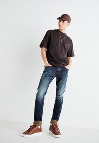 Brun kortärmad t-shirt, urtvättade blå jeans och bruna sneakers med vita sulor. Accessoarer inkluderar ett silverhänge och en brun keps.