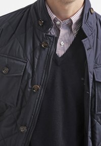 Marineblaue, gesteppte Jacke mit Stehkragen, Knopfdetails und Klappentaschen, getragen über einem schwarzen V-Ausschnitt-Pullover und einem gestreiften Hemd.