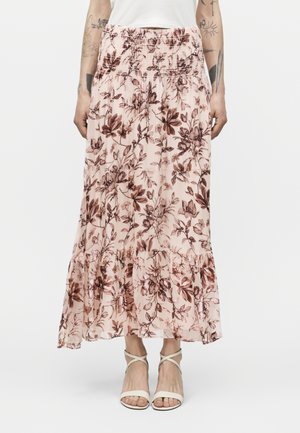 Kobieta w różowej kwiecistej spódnicy midi z elastycznym paskiem i białych sandałach na ramiączkach na jednolitym tle.