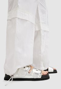 Metallic zilveren sandalen met gestructureerde banden en gespen, geplaatst tegen losse, witte, gekreukte stoffen broeken, die het ontwerp van de schoenen onthullen.