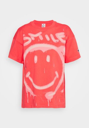 Czerwony t-shirt z krótkim rękawem z dużą różową buźką i słowem "SMILE" nadrukowanym powyżej w stylu cieknącej farby.