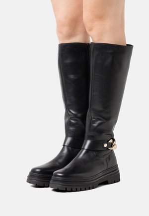 Bottes montantes noires en cuir avec une finition mate, semelle en caoutchouc plate, bride à la cheville ornée d'un accessoire en métal doré. Fermeture zippée latérale.