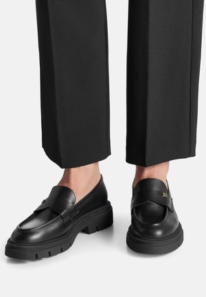 Mocasines de cuero negro con suela gruesa, con una correa frontal y herrajes dorados. Combinados con pantalones negros a medida, que resaltan un diseño elegante.