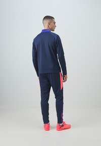 Marineblauer Sportanzug mit lila Akzenten, bestehend aus langärmligem Oberteil und schmal zulaufenden Hosen. Helle pinke Sportschuhe runden das Outfit ab.