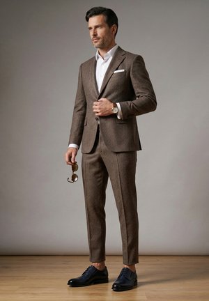 Mand justerer knap på brun jakkesætjakke, iført hvid skjorte, holder solbriller og sorte pæne sko, står på trægulv.