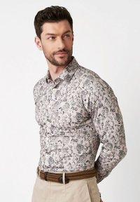 Witte button-up shirt met een zwart bloemenpatroon, lange mouwen en een kraag. Draagt een bruine riem en lichte broek.