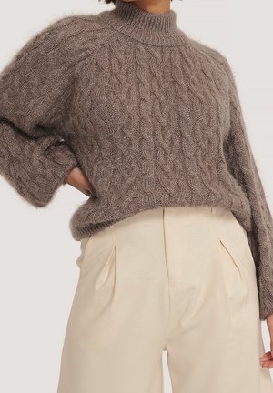 Brązowy, puszysty sweter z grubym splotem i wysokim kołnierzem, włożony do kremowych spodni z wysokim stanem i plisami, zgięty w łokciu przy talii.