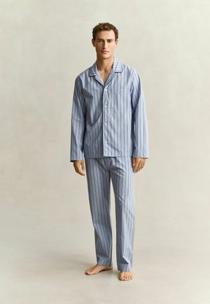 Homme debout pieds nus portant un haut de pyjama à rayures bleu clair, à manches longues, boutonné, et un pantalon assorti sur un fond neutre.