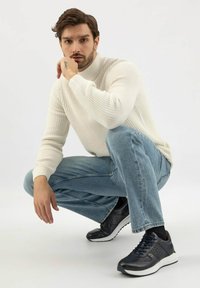 Muž s tmavými vlasy, který si klečí, má na sobě bílý pletený svetr, modré džíny, černé tenisky a opírá bradu o ruku.