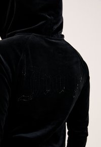 Μαύρο βελούδινο φούτερ με απαλή υφή. Η λέξη "Juicy" εμφανίζεται με μαύρα στρας στην πλάτη, προσθέτοντας μια διακριτική λεπτομέρεια.