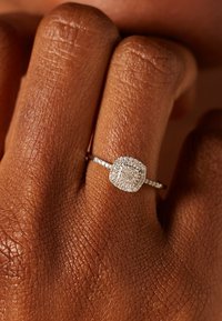 Een ring met diamantenaccenten in zilveren metaal, met een kussenvormig middenstuk dat voorzien is van talrijke kleine diamanten, gezet op een dunne band.