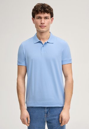 Junger Mann mit kurzen, lockigen braunen Haaren, der ein hellblaues kurzärmeliges Polohemd und blaue Jeans trägt, vor einem schlichten Hintergrund stehend.