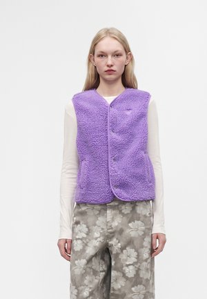 Junge Frau trägt eine violette Fleeceweste über einem weißen Langarmshirt und graue, geblümte, weit geschnittene Hosen, stehend vor weißem Hintergrund.