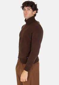 Maglione a collo alto marrone in una texture a coste, design aderente, con maniche lunghe e orlo che si restringe in vita; indossato con pantaloni marroni.