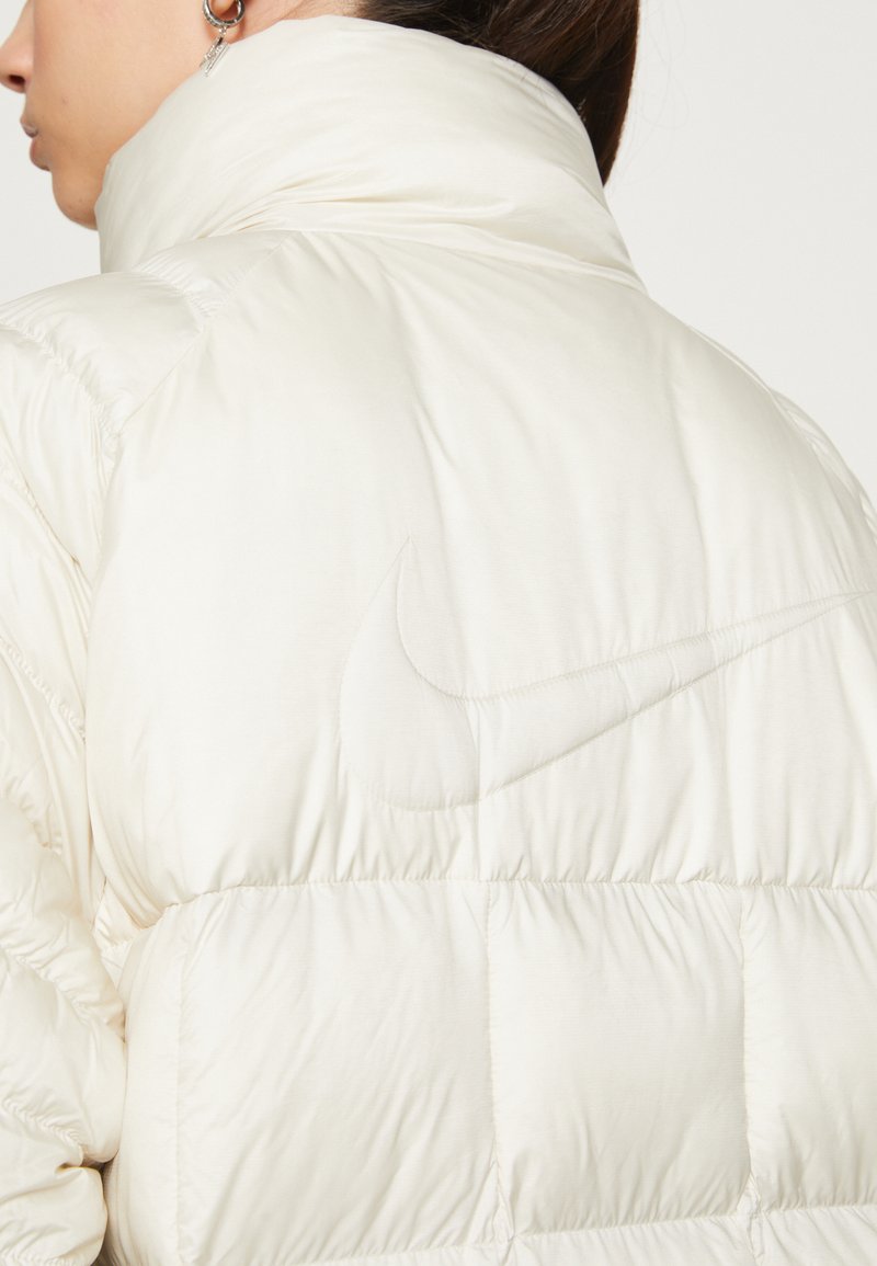 Veste Nike blanche rembourrée avec un col haut, comportant des sections matelassées et un logo Nike ton sur ton subtilement embossé sur le tissu.