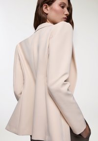 Blazer beige chiaro con spalle strutturate, vestibilità sartoriale e tessuto liscio. Nessun hardware visibile; presenta una singola fessura sulla parte posteriore.