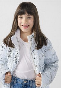 Chaqueta azul claro con lentejuelas, cierre frontal con cremallera, puños acanalados y cuello alto, llevada sobre una camiseta blanca y vaqueros azules.