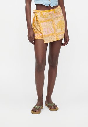 Personne portant un haut court bleu clair, une jupe portefeuille jaune à motifs tropicaux et des tongs vertes, debout sur un fond blanc.