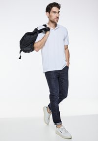 Bílá polo tričko, tmavé džíny a bílé tenisky. Nese černý batoh, pózuje na světlém pozadí. Úhledný a neformální outfit.