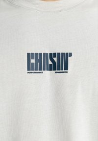 Camiseta de color gris claro con un gráfico atrevido en azul oscuro que dice "CHASIN' PERFORMANCE JEANSWEAR". Fabricada con material de algodón suave.