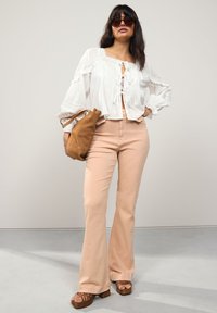 Biała bluzka z falbankami na rękawach, wiązana przy szyi, zestawiona z jasnoróżowymi rozkloszowanymi spodniami. Brązowa skórzana torebka i sandały na platformie dopełniają stylizację.