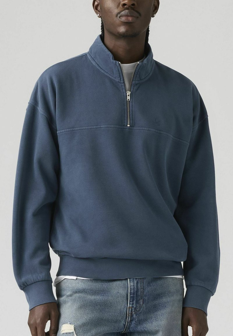 Hombre con sudadera azul con cremallera hasta el pecho y jeans azules claros desgastados, presentado sobre un fondo liso.