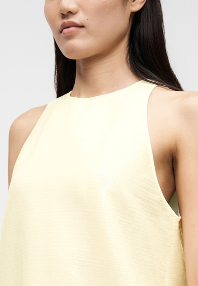Ανοιχτό κίτρινο αμάνικο τοπ με λείο, υφασμάτινο ύφασμα με υφή. Διαθέτει ψηλό λαιμό και φαρδιά μασχάλες, προσφέροντας μια ά νετη εφαρμογή.