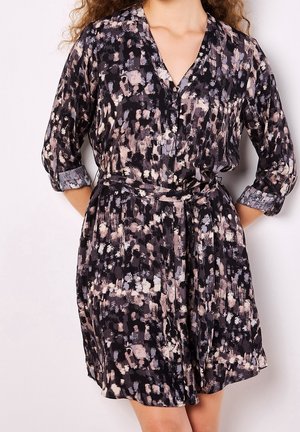 Kobieta ubrana w sukienkę zapinaną na guziki, z długim rękawem i paskiem, z abstrakcyjnym wzorem w kolorach czarnym, beżowym i fioletowym, rękawy podwinięte do łokci.