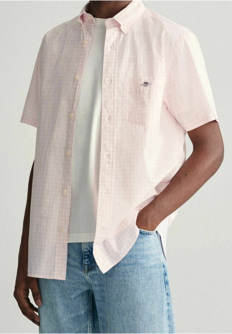 Camisa de botones de manga corta en un patrón de cuadros rosa claro, hecha de algodón, con un bolsillo, collar y corte relajado. Lleva una camiseta blanca debajo.