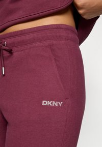 Pantaloni de trening burgundy dintr-un material moale, cu talie ajustabilă cu șnur, buzunare laterale și detaliu cu logo din cristal pe gambă.