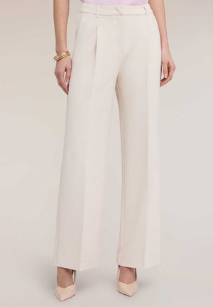 Mujer con pantalones crema de talle alto y pierna ancha, combinados con zapatos de tacón rosa pálido, pulseras doradas y una blusa rosa claro.