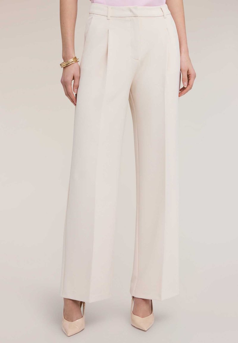 Nainen pukeutuneena korkeavyötäröisiin, leveälahkeisiin kermaan vivahtaviin housuihin, joita täydentävät vaaleanpunaiset korkokengät, kultaiset rannekorut ja vaaleanpunainen toppi.