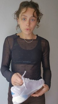 Γυναίκα με κατσαρά μαλλιά κρατάει ένα λευκό αθλητικό παπούτσι, φοράει μαύρο διάφανο τοπ με μακριά μανίκια και ασημένιο αλυσίδα κολιέ.