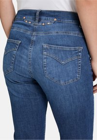 Modré džíny z denimu s polovičním pasem, designem na pět kapes a měděnými akcenty. Obsahují blednutí a přizpůsobené švy na zadních kapsách.